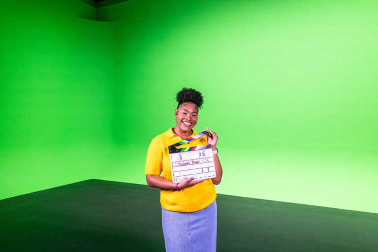 Cheerful young woman in a yellow shirt holding a film clapperboard, standing against a bright green background, representing 'ACC Student Beat'.
