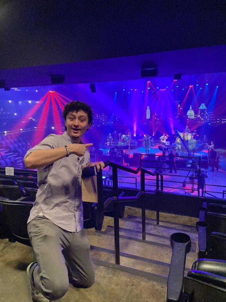 Young student excitedly pointing towards a stage setup with musicians and vibrant lighting, representing ACC's coverage of local Austin events.