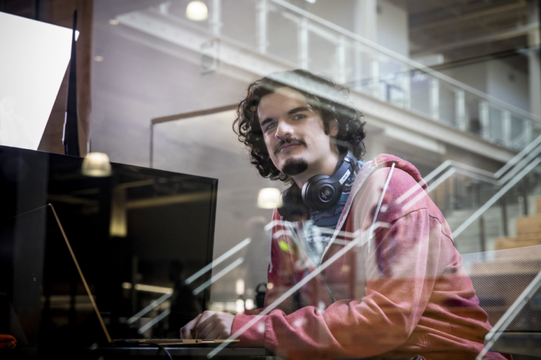 Young man with curly hair and headphones, working intently on a computer in a modern building with stairs and large windows, representing a student featured in 'Day in the Life' docuseries.
