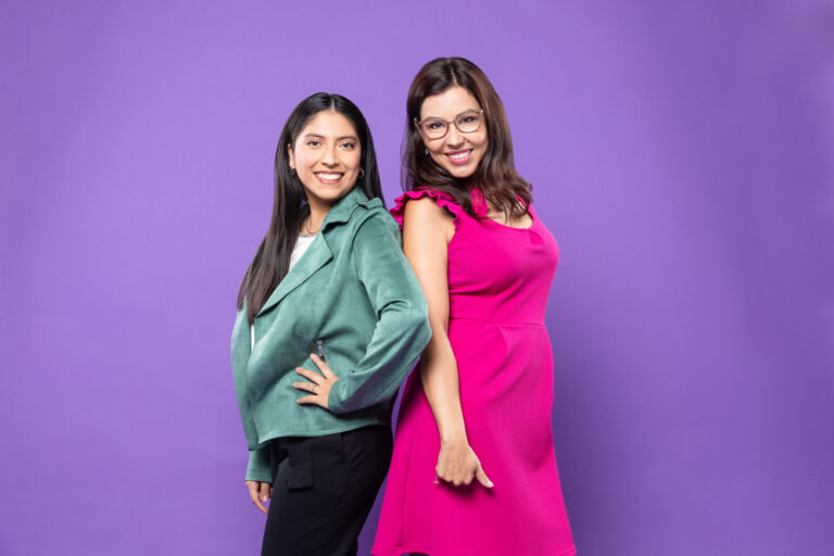 Two confident women, one in a teal jacket and the other in a pink dress, standing back-to-back against a purple background, representing the hosts of 'Ready, Set, Ascender!' talk show.
