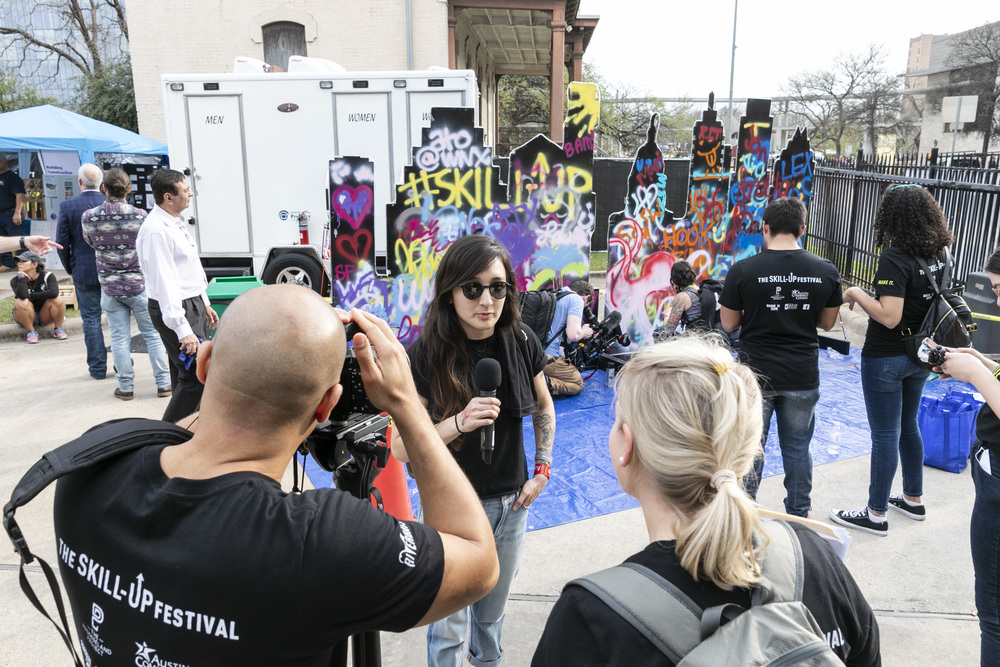 A photographer captures a young woman being interviewed, holding a microphone, at an outdoor event. Behind them are colorful graffiti walls, attendees wearing 'The Skill-Up Festival' shirts, and mobile restroom trailers labeled 'Men' and 'Women'. The scene embodies a lively, community-driven atmosphere.
