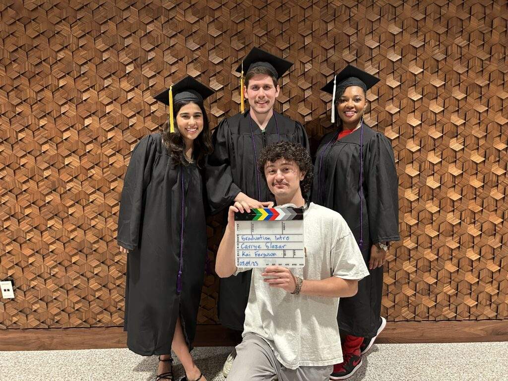 Group of four Austin Community College students in graduation gowns; three standing with caps and one kneeling in front holding a clapperboard, all posing in front of a textured wooden wall.