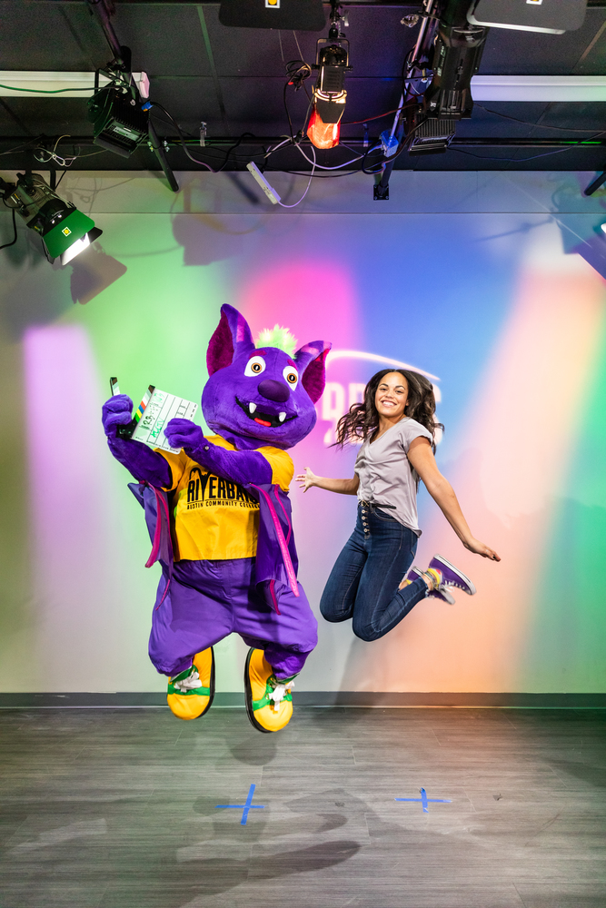 Purple mascot named R.B. holding a film clapperboard while a joyful young girl jumps beside him, set against a colorful background, representing the 'R.B. and Friends' show.