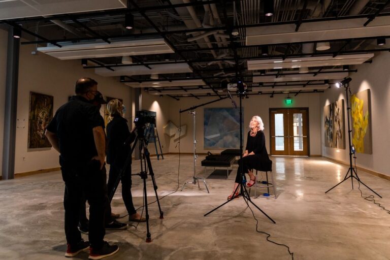 Camera crew setting up to interview a seated woman with curly blonde hair in a contemporary art gallery, representing Austin's vibrant art, music, and entertainment scene.