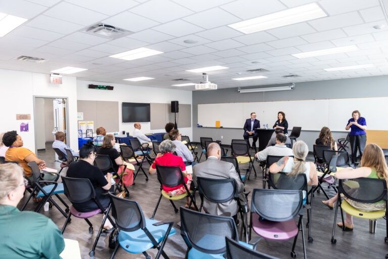 Diverse group of attendees seated in a modern conference room listening intently to two speakers at the front, discussing community and culture topics at an Austin Community College event.